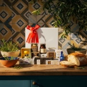 Artisanal Greek food hamper in a rustic kitchen setting. The luxury white gift box with a red bow is on a wooden counter, surrounded by gourmet Greek products, fresh herbs, olive branches, and artisanal bread.