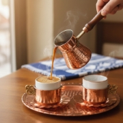 Authentic Greek coffee being poured from a handcrafted copper briki into a porcelain cup on a matching hammered tray, showing rich kaimaki foam.