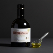 Luxury dark glass bottle of Elea’s Poema Ode 1 next to a glass apothecary spoon filled with high-polyphenol green olive oil against a dark charcoal background.